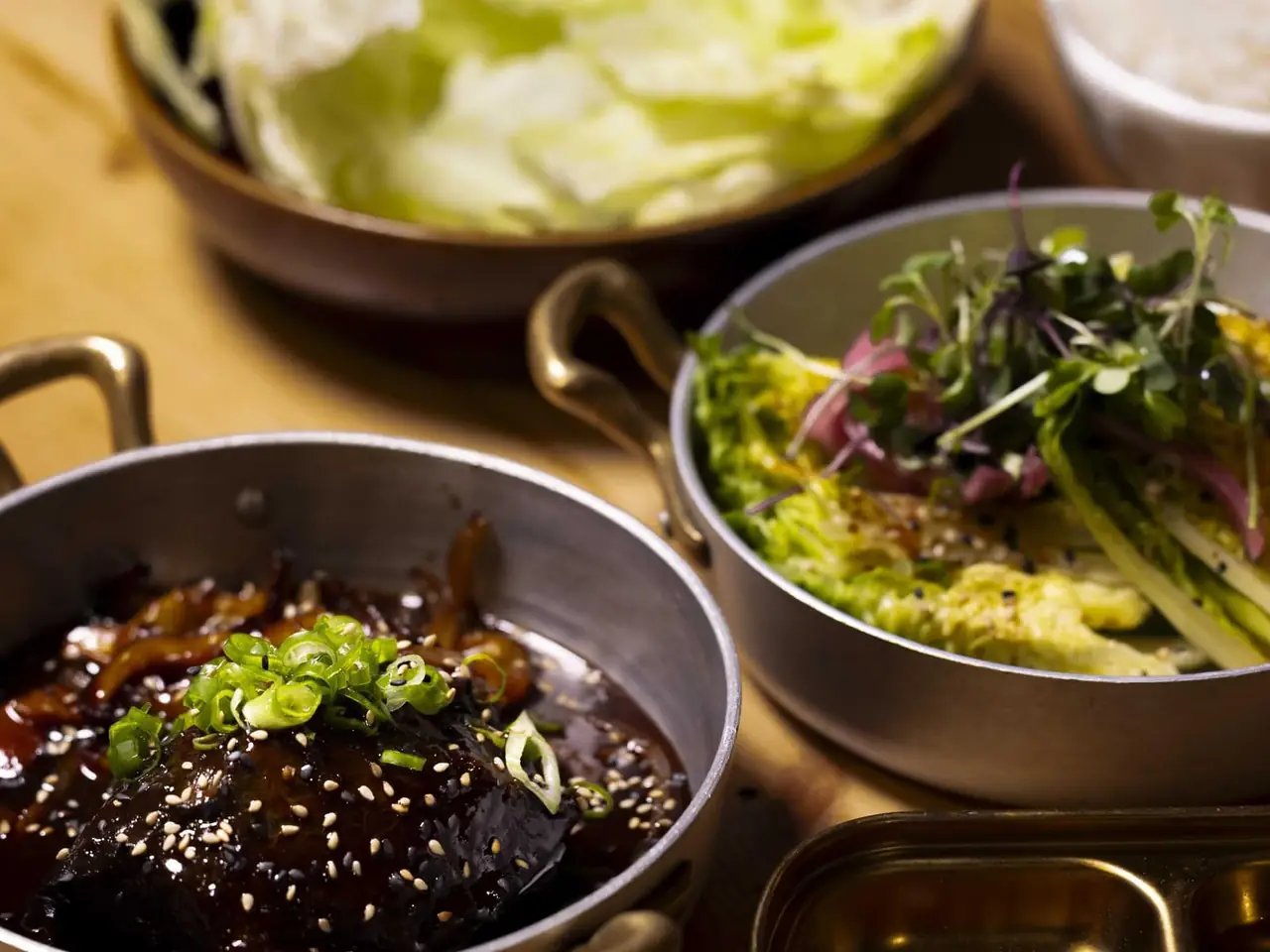 Two metal pans of Korean dishes on a wooden table — braised meat glazed in dark sauce topped with scallions and sesame seeds, and a slaw-like dish with pickled red onion and microgreens, San Kil