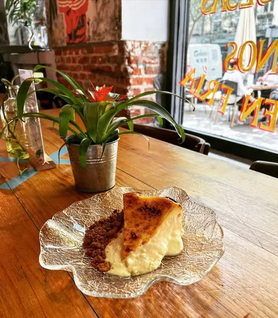 Slice of caramelized cheesecake or torrija with crumb on a textured glass plate, beside a potted plant on a wooden table by a window