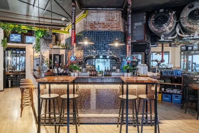 Central bar with a wood-topped counter and stools, blue-tiled backsplash, beer taps and hanging bottles, with brewing tanks overhead