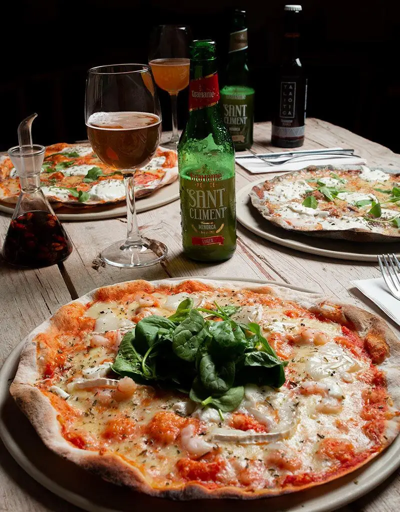 Wood-fired pizza topped with fresh spinach leaves, mozzarella, and shrimp, served on a wooden table with beer and wine at Santa Clara
