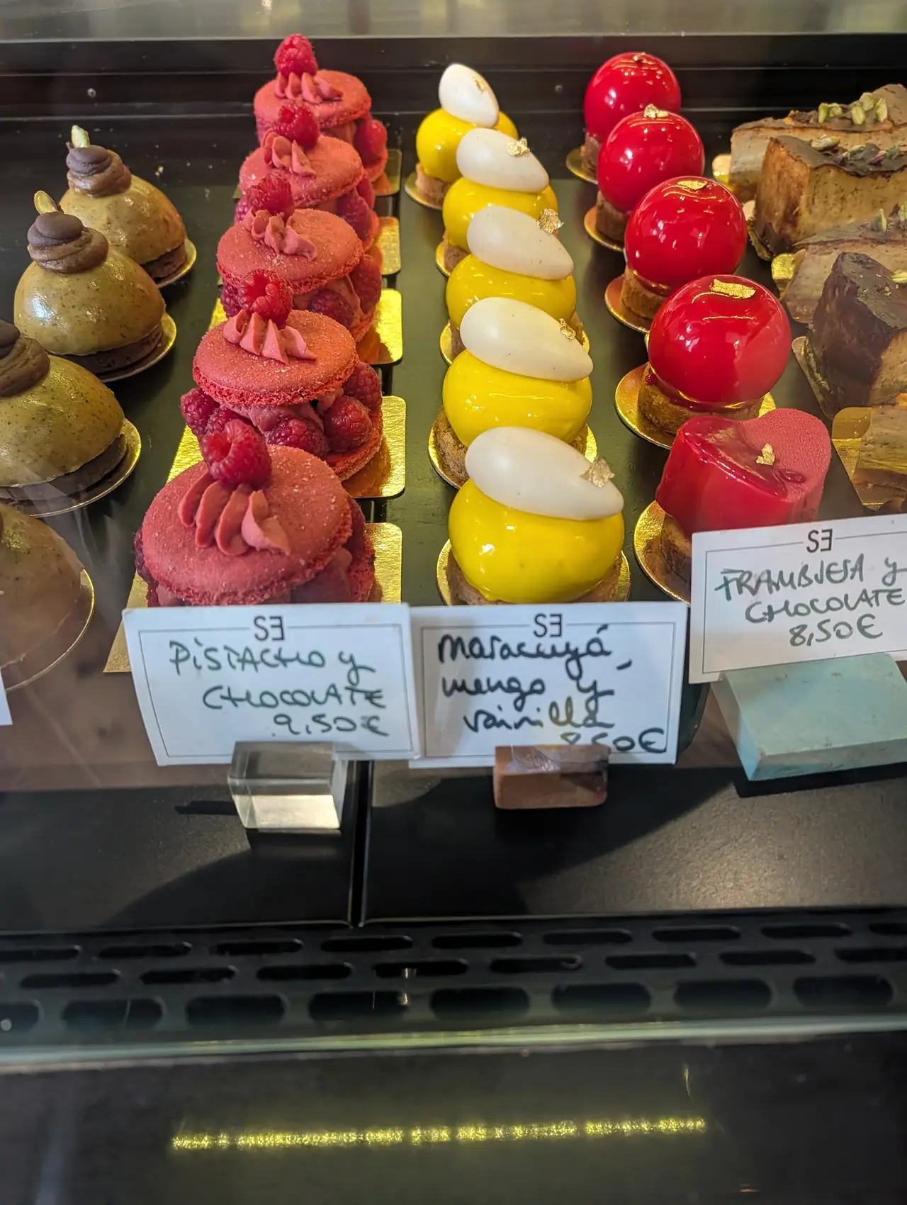 Display case of individual pastries including pistachio-chocolate domes, raspberry macaron towers and mango-passionfruit mousse cakes