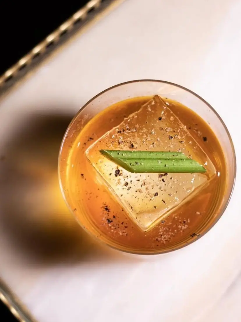 Overhead view of an amber cocktail over a large clear ice cube with green celery garnish on a marble surface