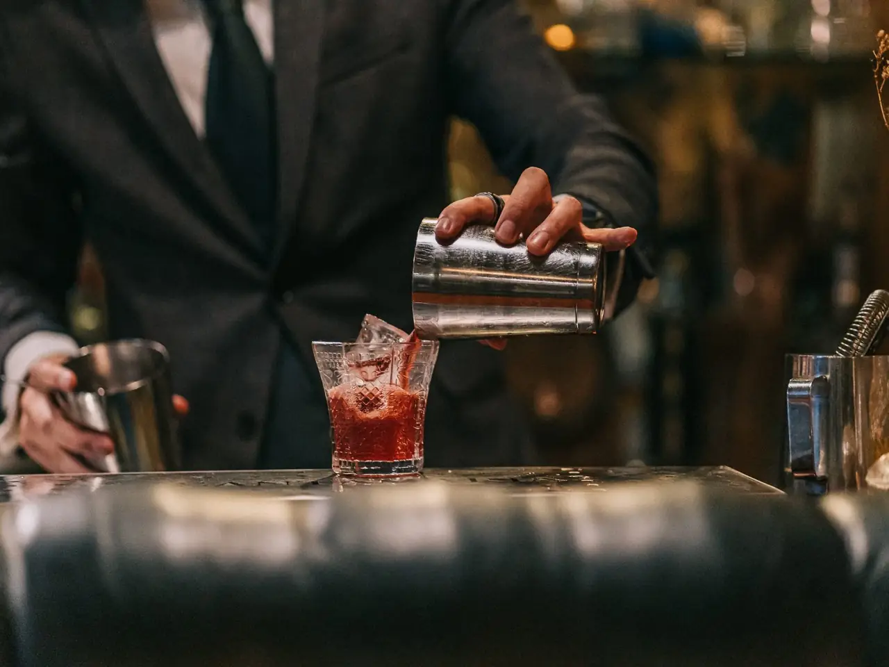 Bartender in a suit pouring a red negroni-style cocktail from a metal shaker into a cut-crystal glass at the bar