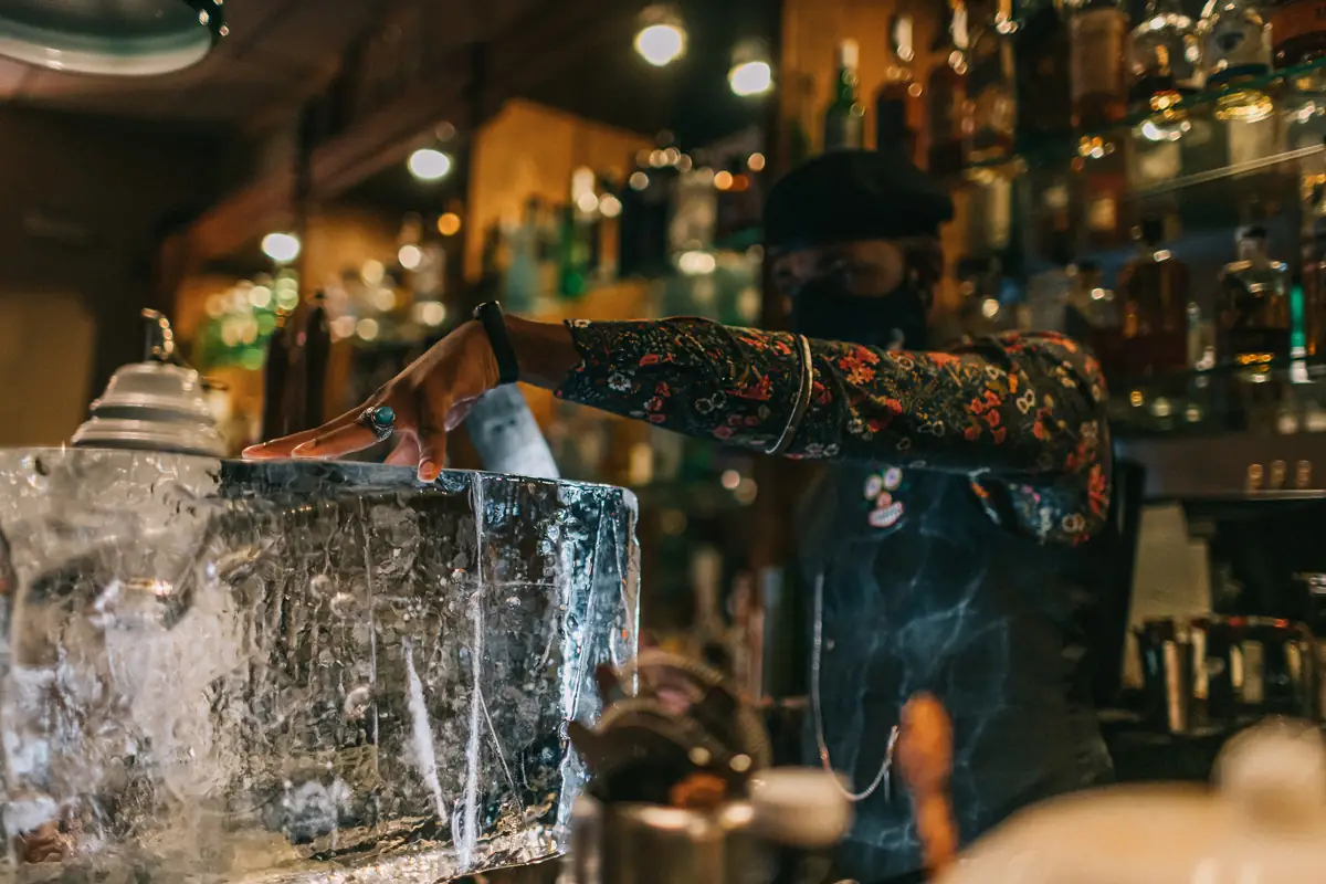 Bartender reaching over a large clear ice block behind a dimly lit cocktail bar lined with spirit bottles