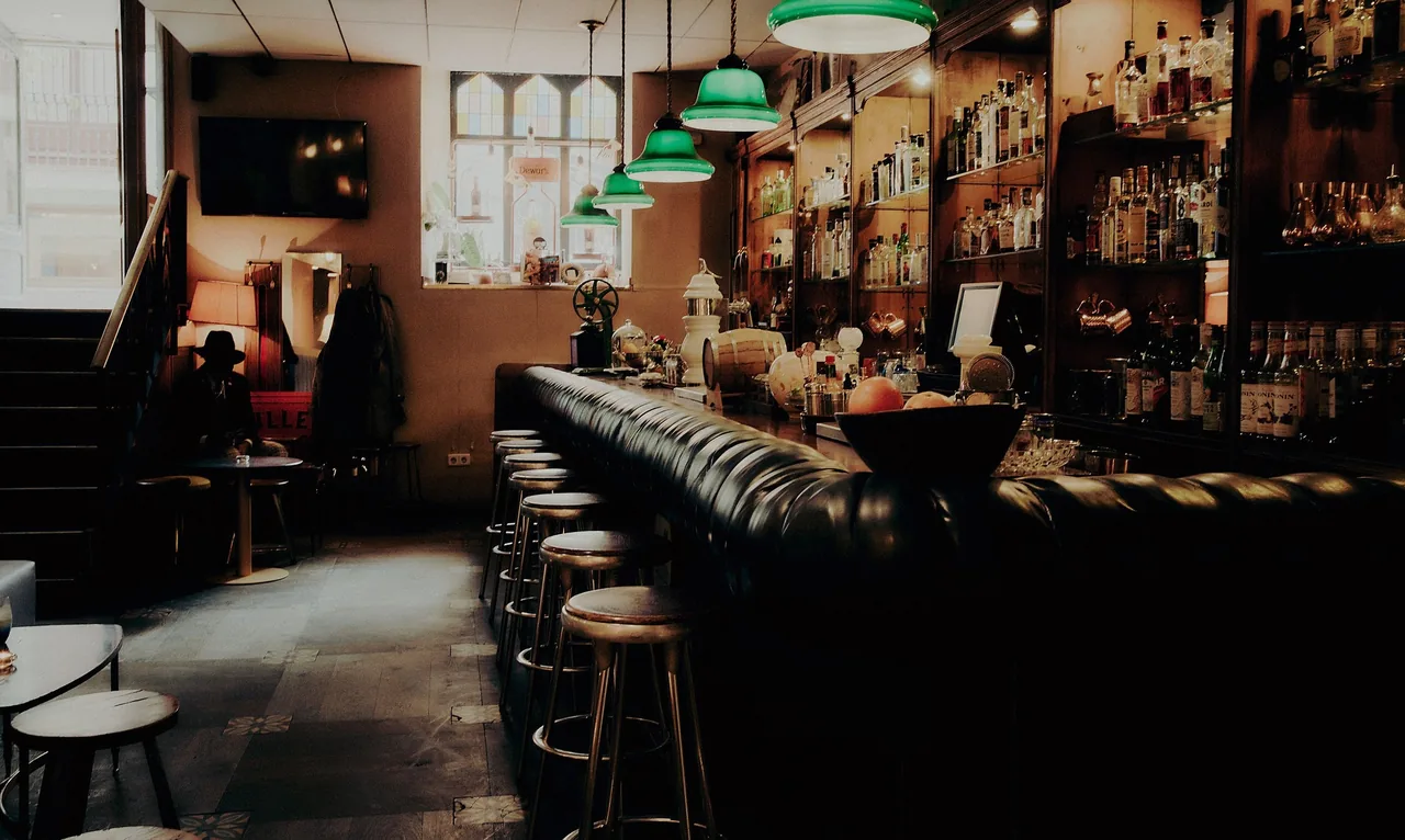 A bar counter with a row of stools, green pendant lamps and shelves of bottles