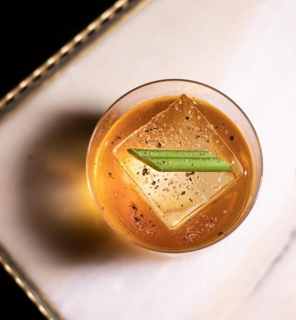 A cocktail with a large ice cube and a green garnish seen from above on a white surface