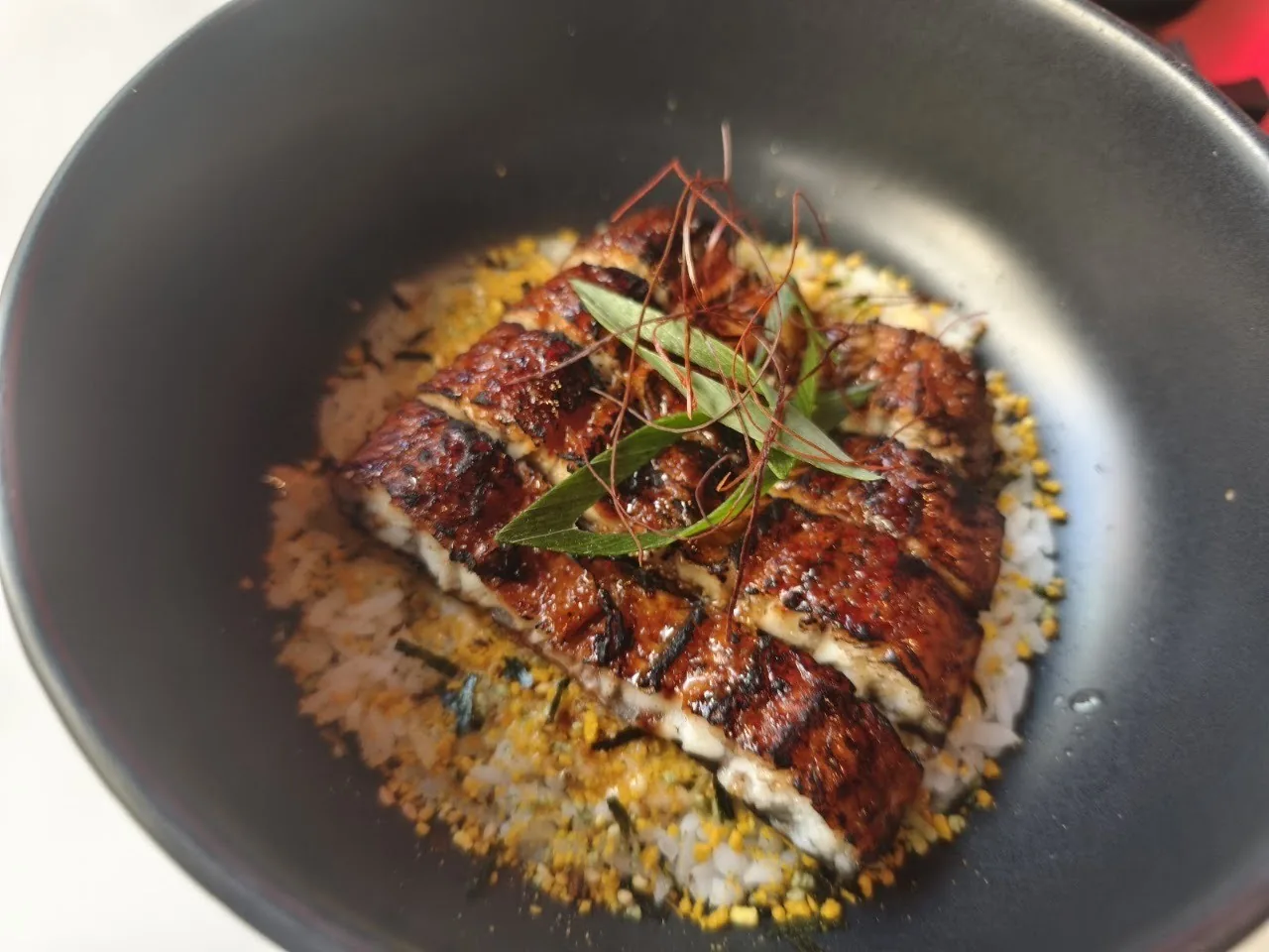 A dark bowl of glazed grilled eel over rice with herbs