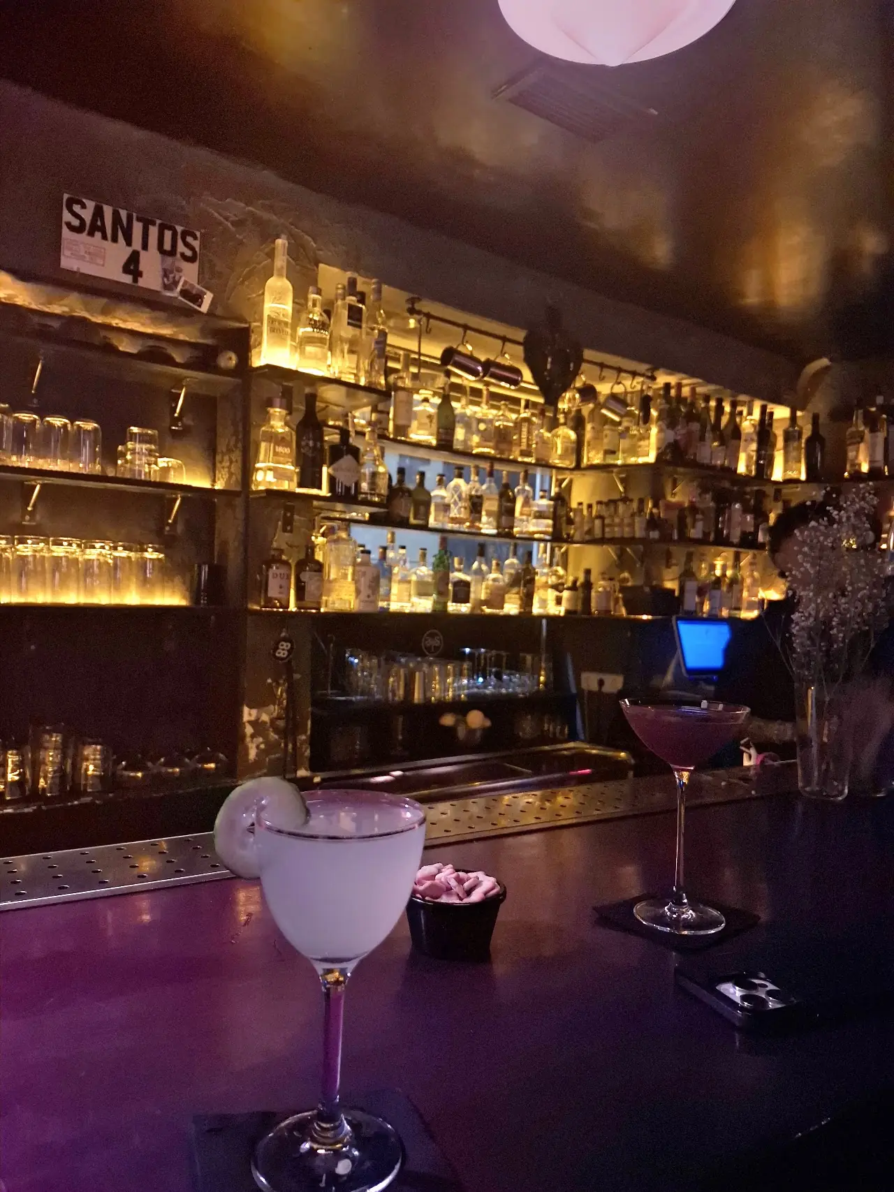 Two cocktails on the bar in front of backlit shelves of bottles and the Santos sign