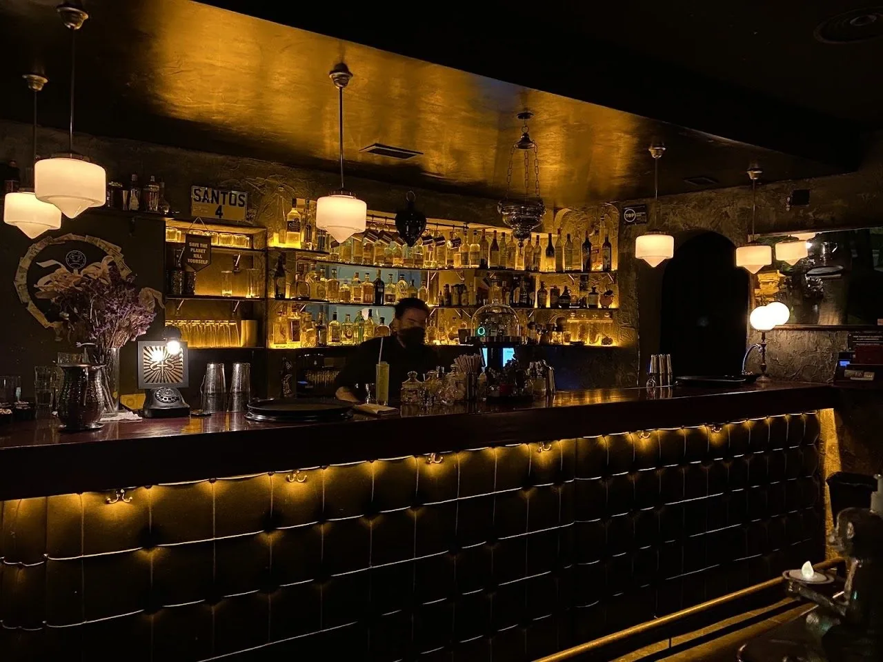 A long bar with padded front, hanging lamps and shelves of bottles behind it