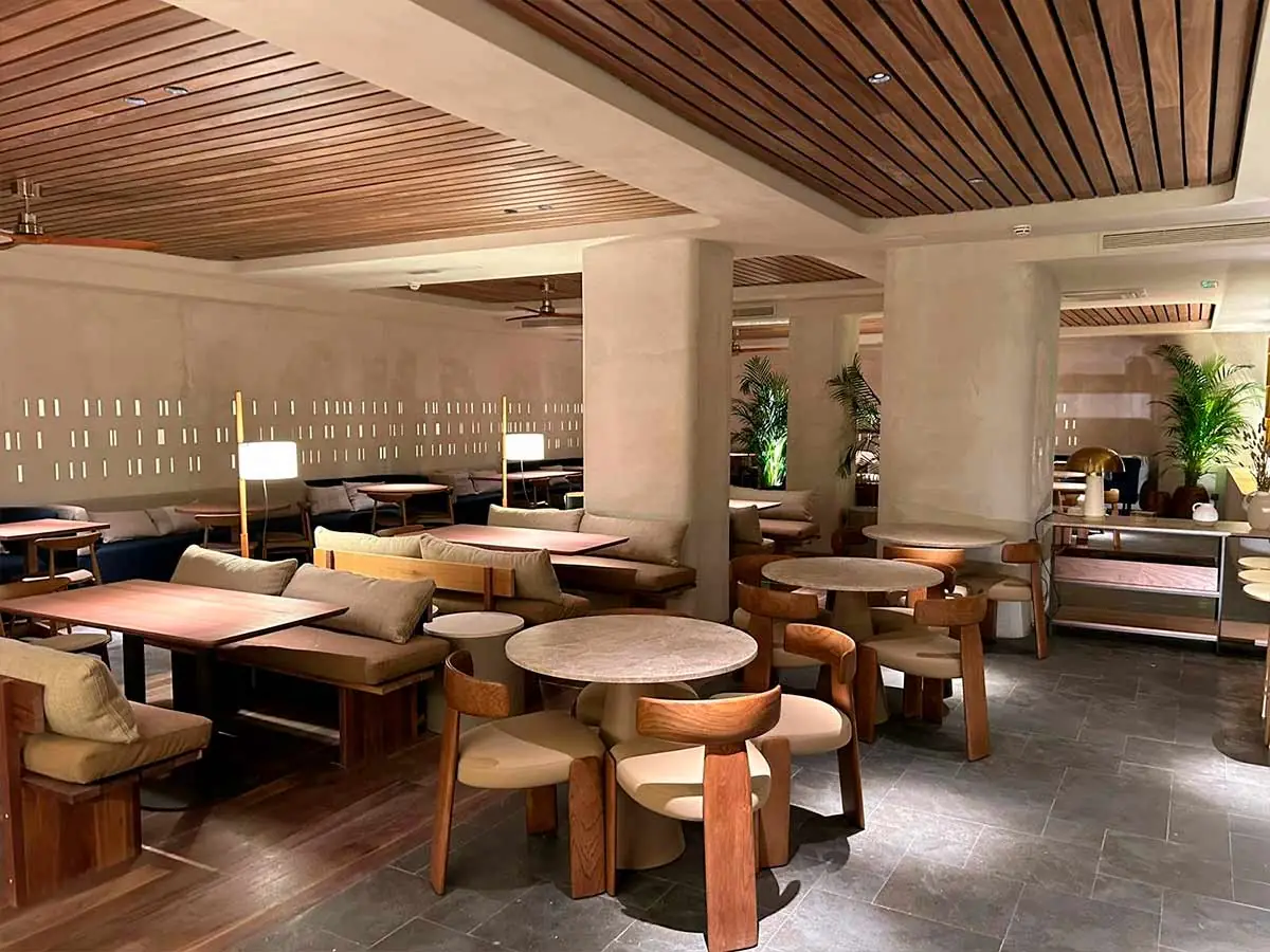Warm-toned restaurant dining room with wood-slat ceiling, cushioned banquettes, round travertine tables and pale wood chairs on grey stone flooring
