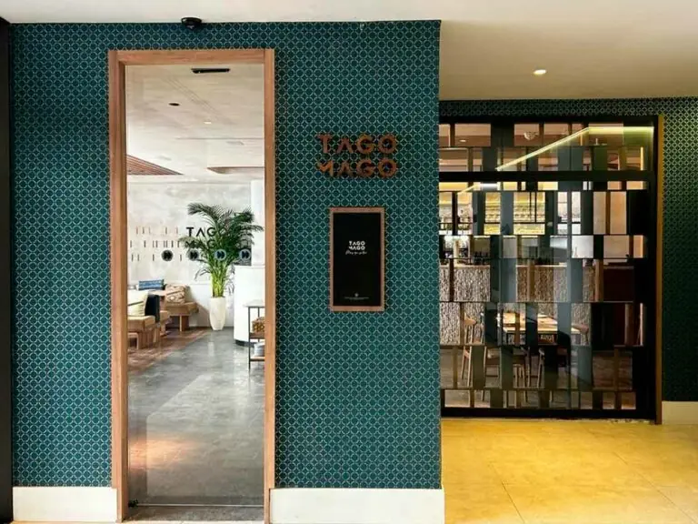 Restaurant entrance with green patterned tile wall and copper 'Tago Mago' lettering beside a glass door onto the dining room
