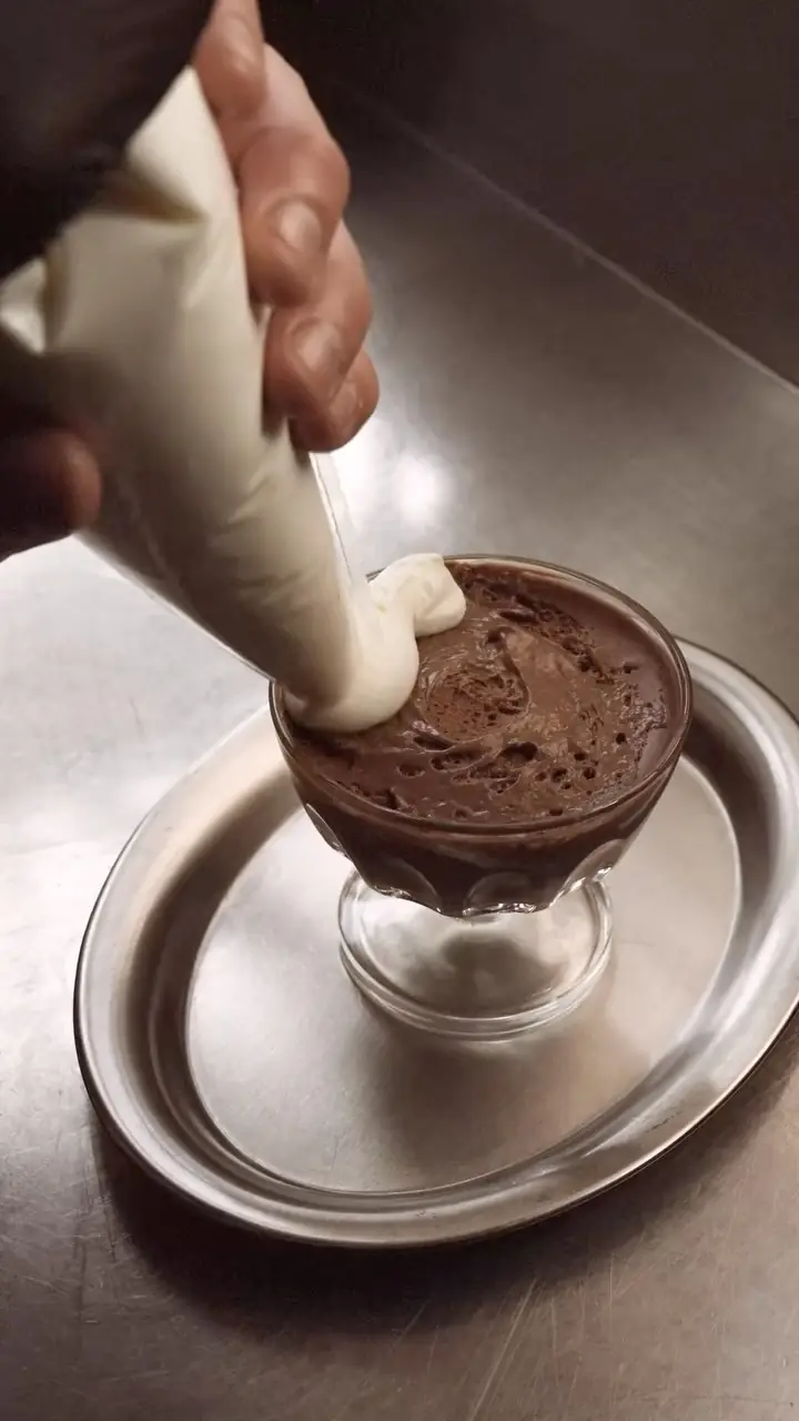 A chocolate mousse dessert in a glass cup being finished with a piped cream topping