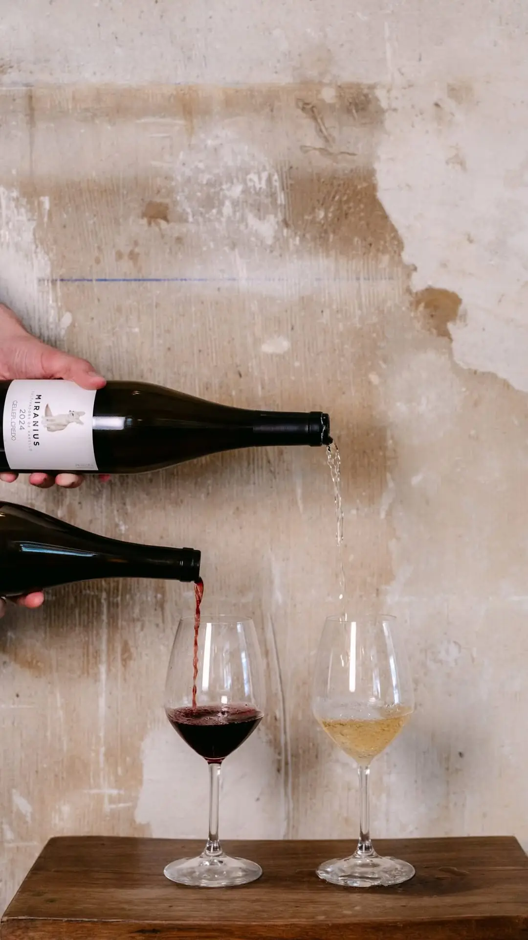 Two hands pouring red and white wine into glasses against a weathered wall
