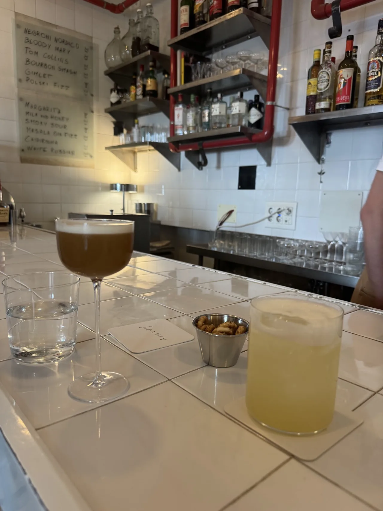 A tiled bar counter with a coupe cocktail and a yellow drink in front of shelves of bottles