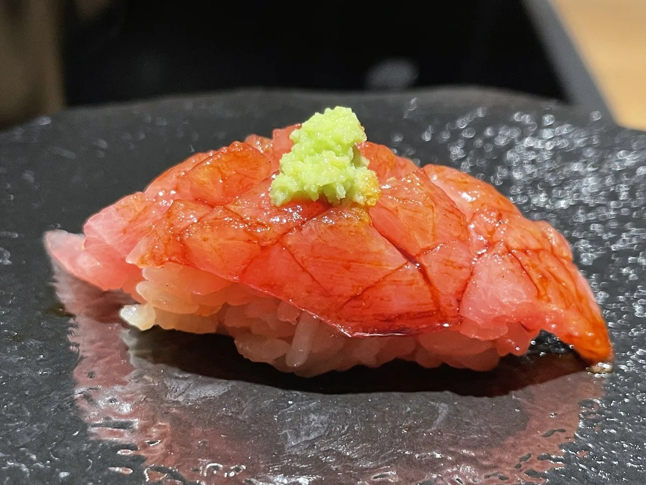 A piece of tuna nigiri topped with a dab of wasabi on a dark stone plate