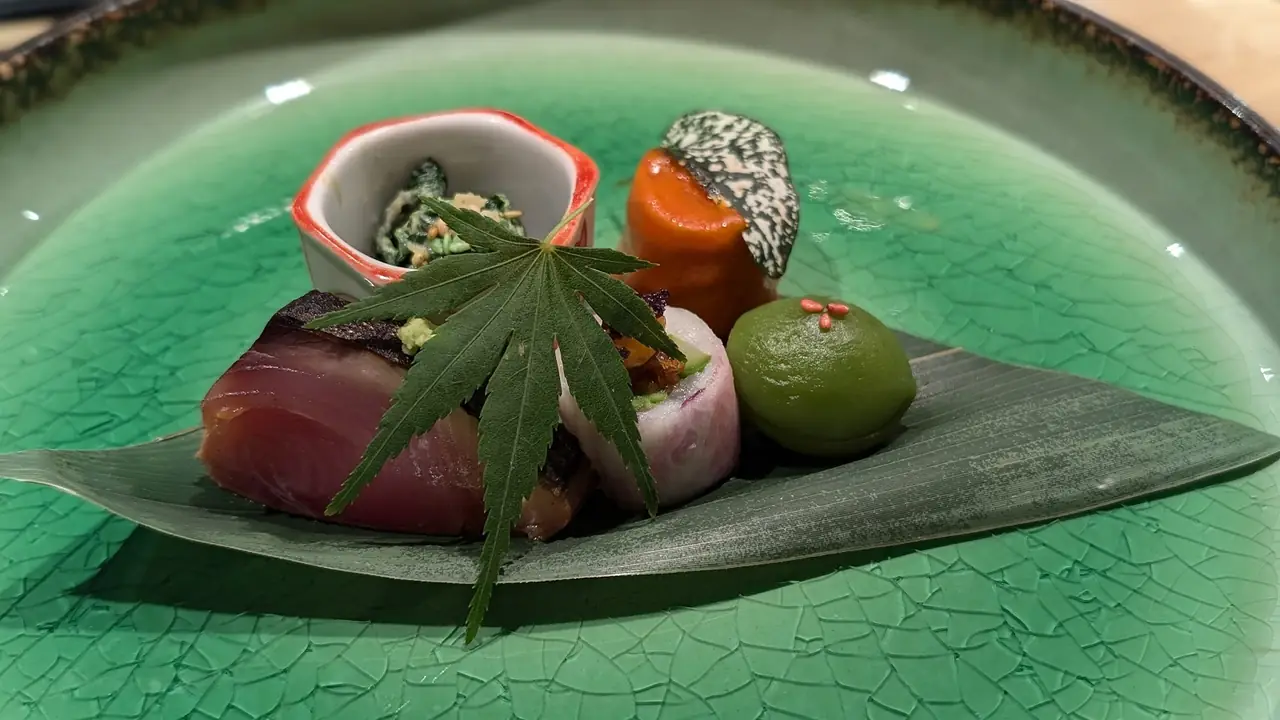 Sashimi and small bites plated on a bamboo leaf with a maple leaf garnish on a crackle-glazed green plate