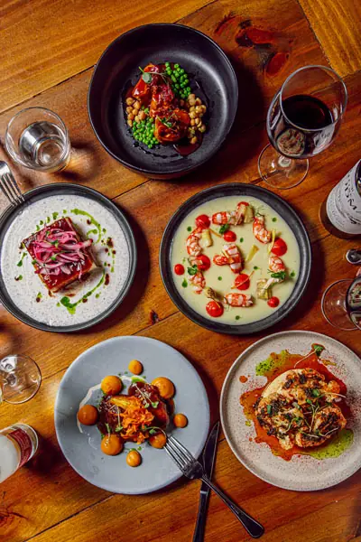 Overhead view of five tapas plates on a wooden table, including a slab of glazed pork belly with pickled onions, prawns in a light sauce, roasted peas with tomato, and other small plates with wine — Sensi Tapas