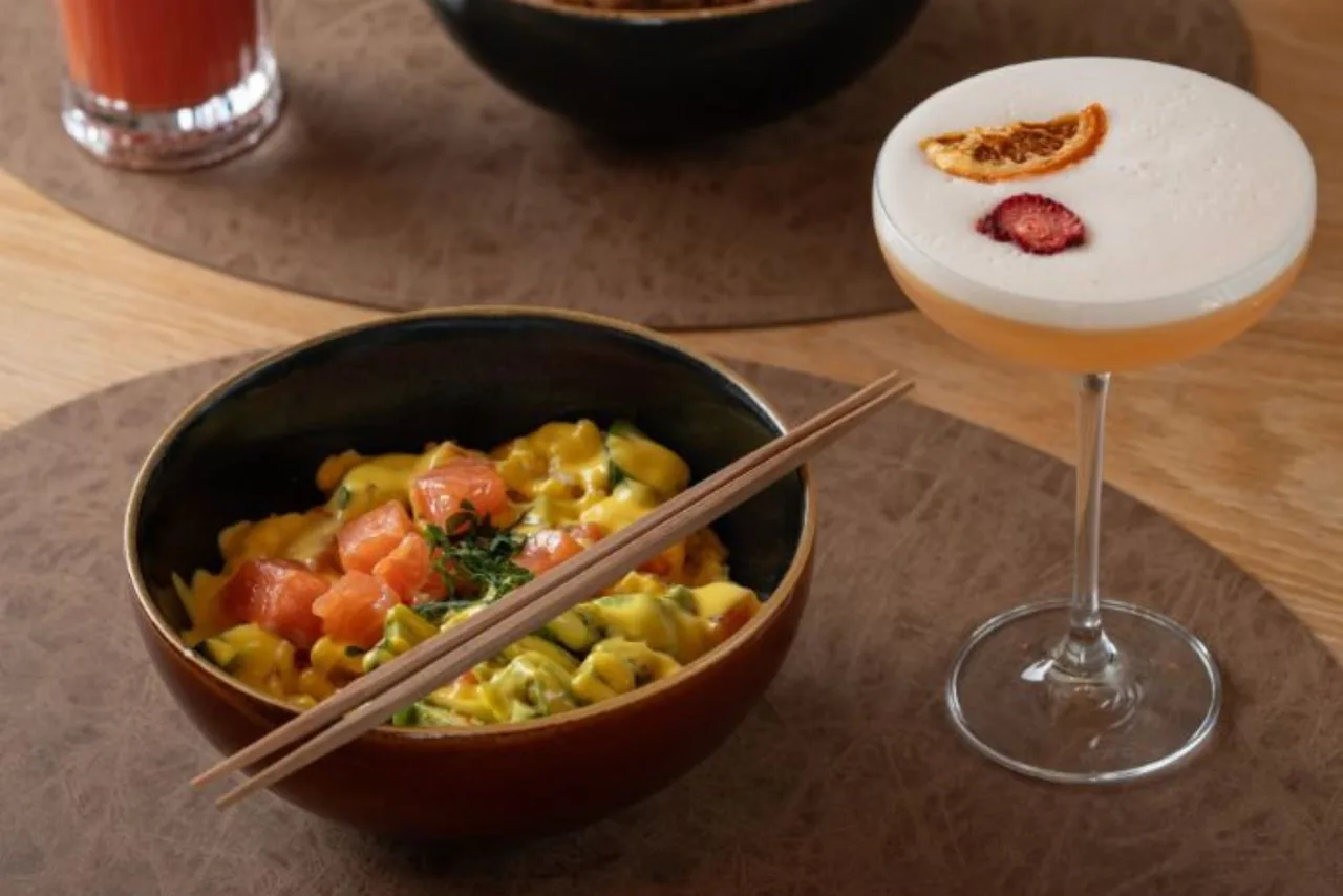 A bowl of diced food beside a frothy cocktail in a coupe glass on a wooden table
