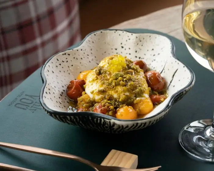 A bowl of burrata with cherry tomatoes and pistachio crumble on a table, with a glass of white wine beside it