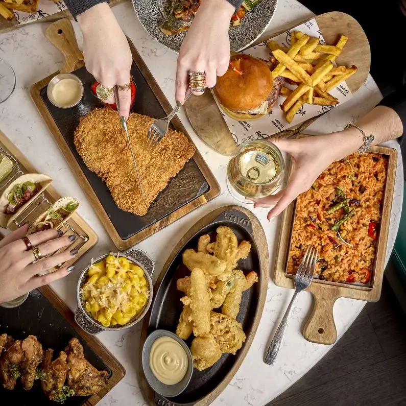 Overhead table spread of breaded schnitzel, a burger with fries, tomato rice, mac and cheese, tempura, small tacos and a glass of white wine with diners' hands reaching in