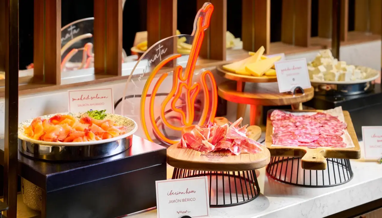 Buffet display with a platter of smoked salmon, a board of sliced jamon iberico, a board of salchichon and stands of cheese, beside an orange neon guitar sign