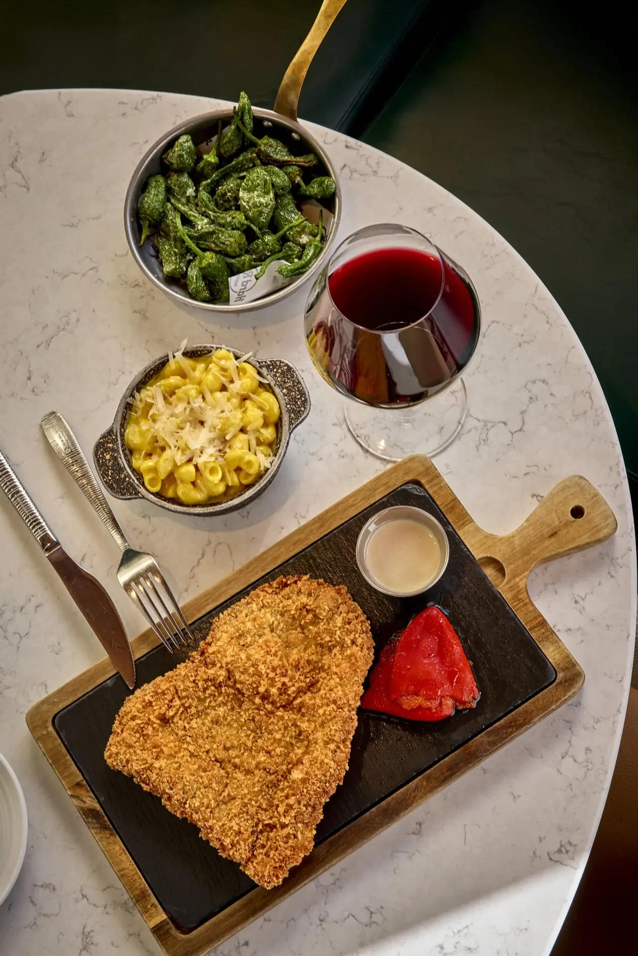 Overhead of a breaded schnitzel with a roasted red pepper and creamy sauce on a slate board, a pot of mac and cheese, a bowl of blistered padron peppers and a glass of red wine
