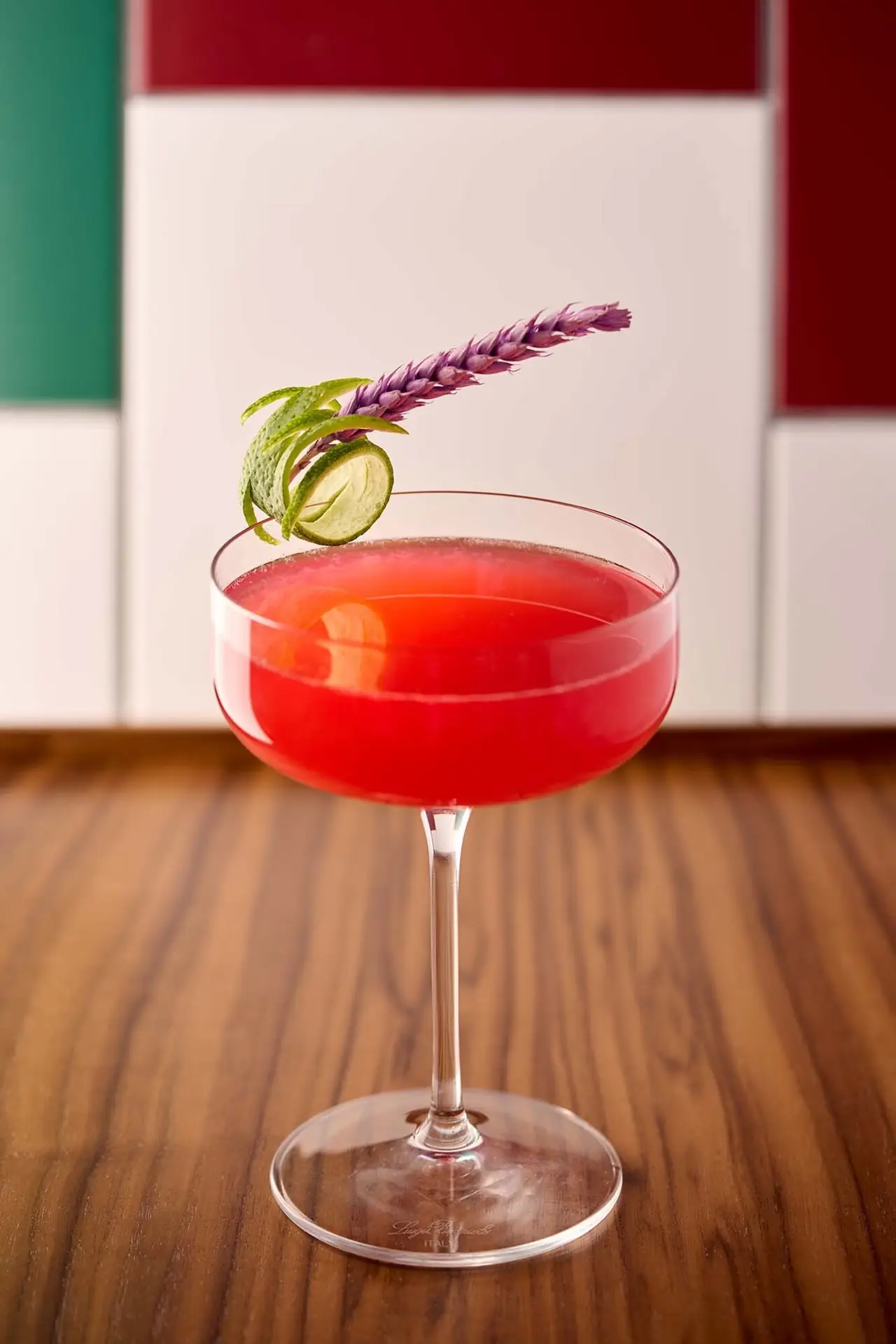 Bright red cocktail in a coupe glass garnished with a coiled lime peel and a lavender sprig, on a wood surface against a green and red tiled wall