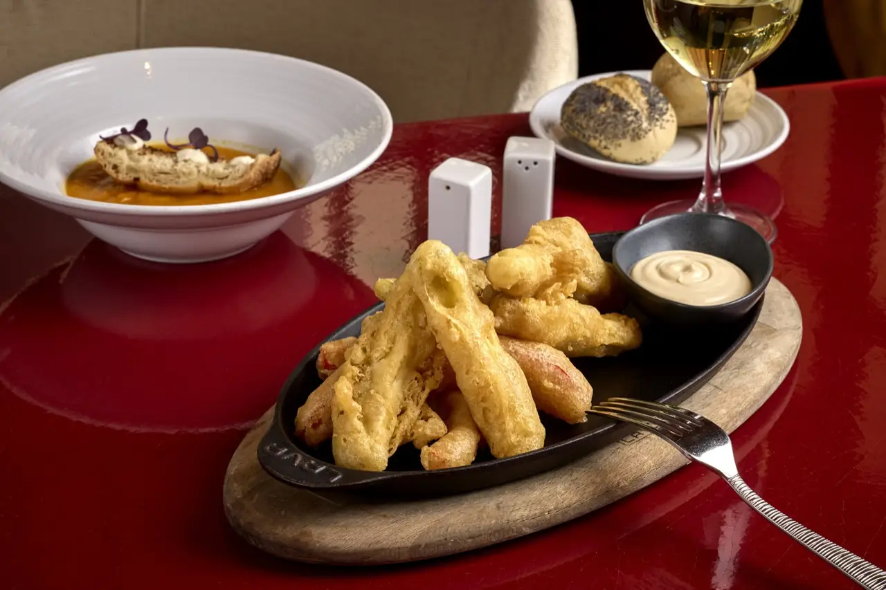 Oval cast-iron dish of tempura prawns with a pot of aioli, a bowl of orange soup with toasted bread, a poppy-seed roll and a glass of white wine on a red table