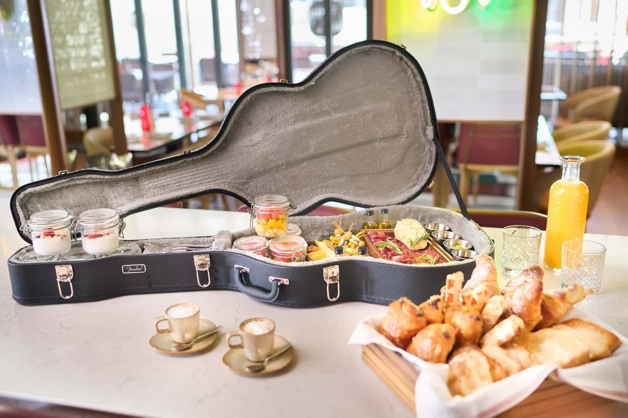 An open guitar case holding jars of yoghurt and fruit and a charcuterie board, beside a basket of croissants, two coffees and a jug of orange juice