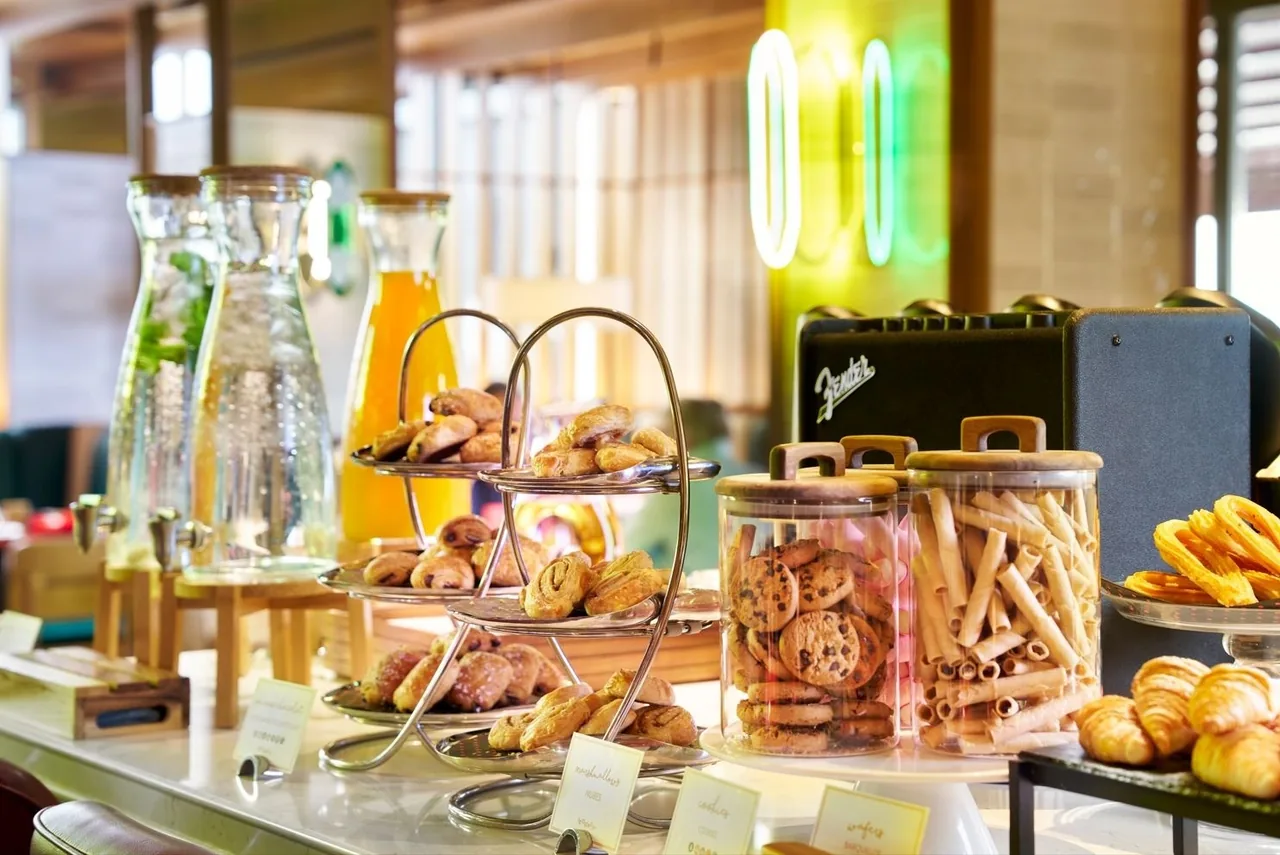 A breakfast buffet counter with tiered stands of small pastries, jars of cookies, croissants and carafes of juice and water