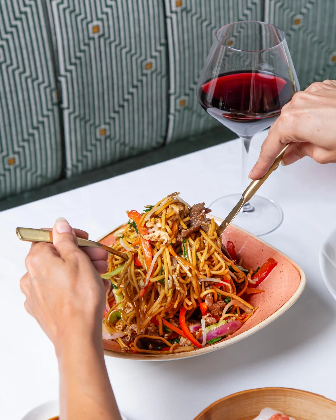 Bowl of stir-fried lo mein noodles with beef, peppers, and bean sprouts, served next to a glass of red wine