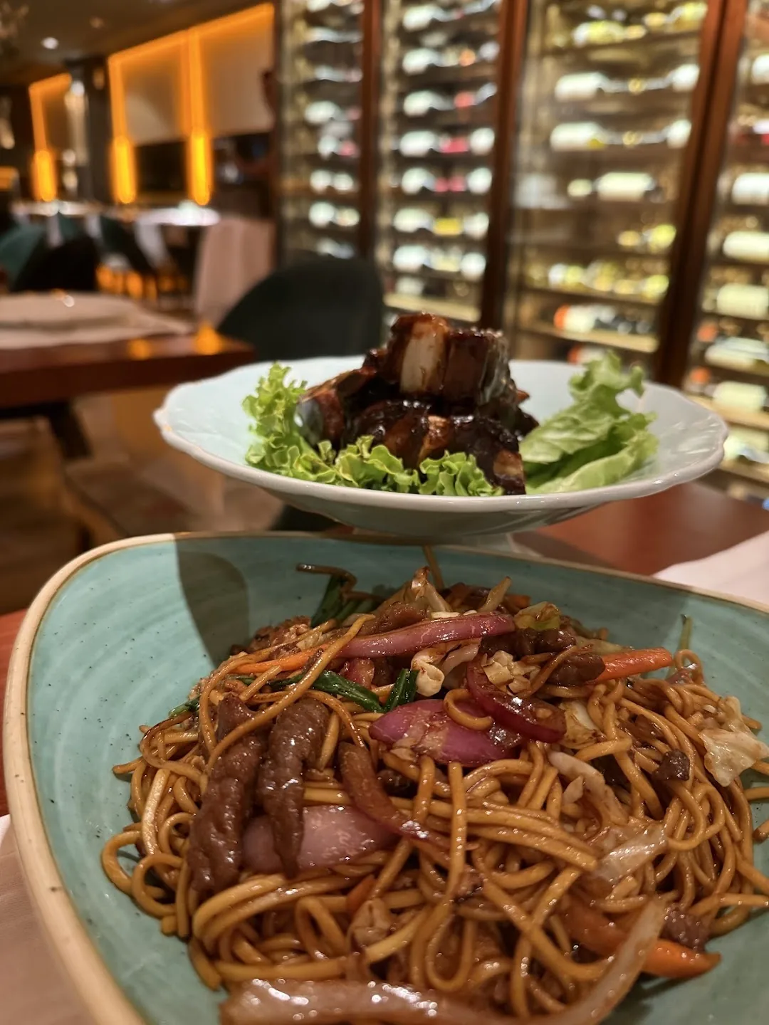Two plates: glazed pork belly on lettuce and stir-fried noodles with beef and red onion