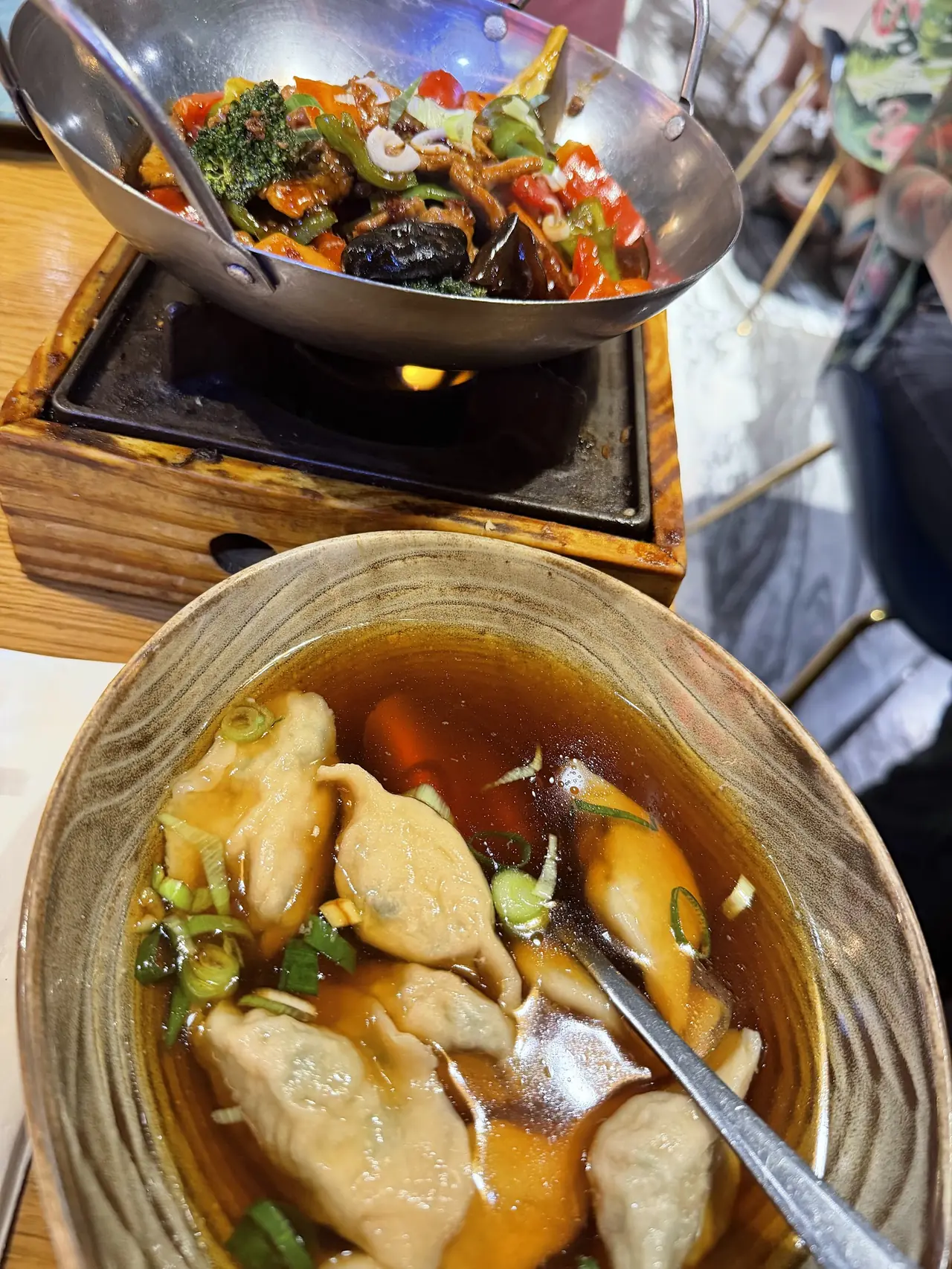 Stir-fried vegetables in a wok over a tabletop burner, with a bowl of dumplings in broth in the foreground