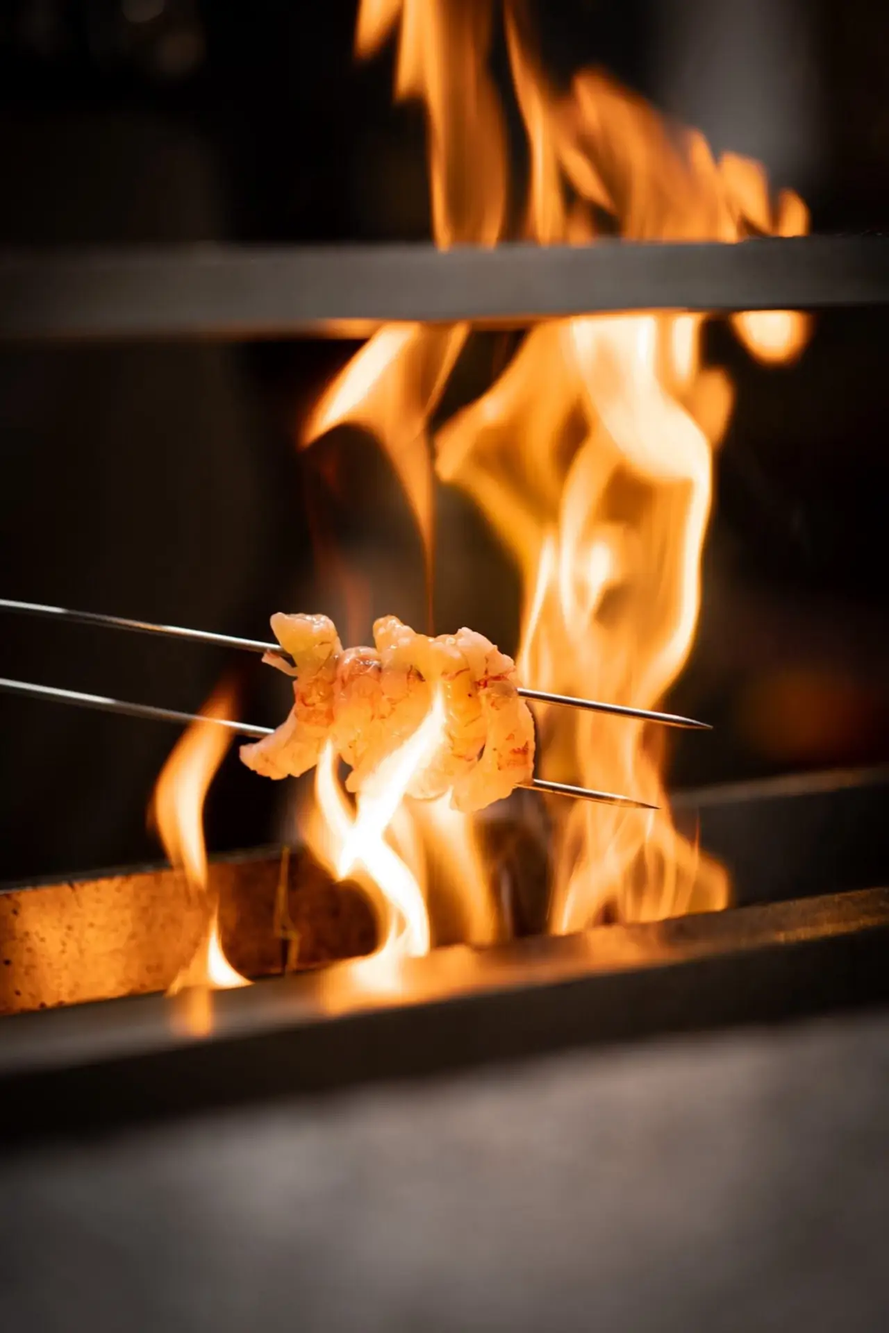 Prawns on twin metal skewers grilling over open orange flames on a charcoal grill