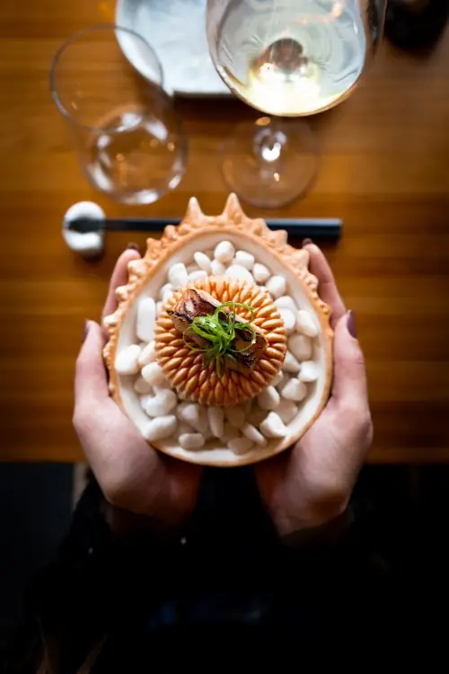 Hands holding a decorative shell-shaped plate of a scallop dish set among white pebbles, topped with shaved scallion
