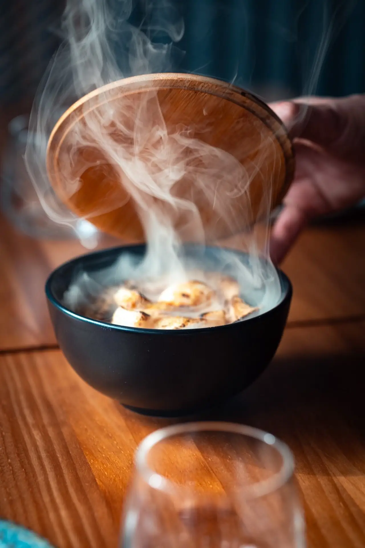 Black bowl of grilled morsels releasing white smoke as a wooden lid is lifted off, on a wooden table