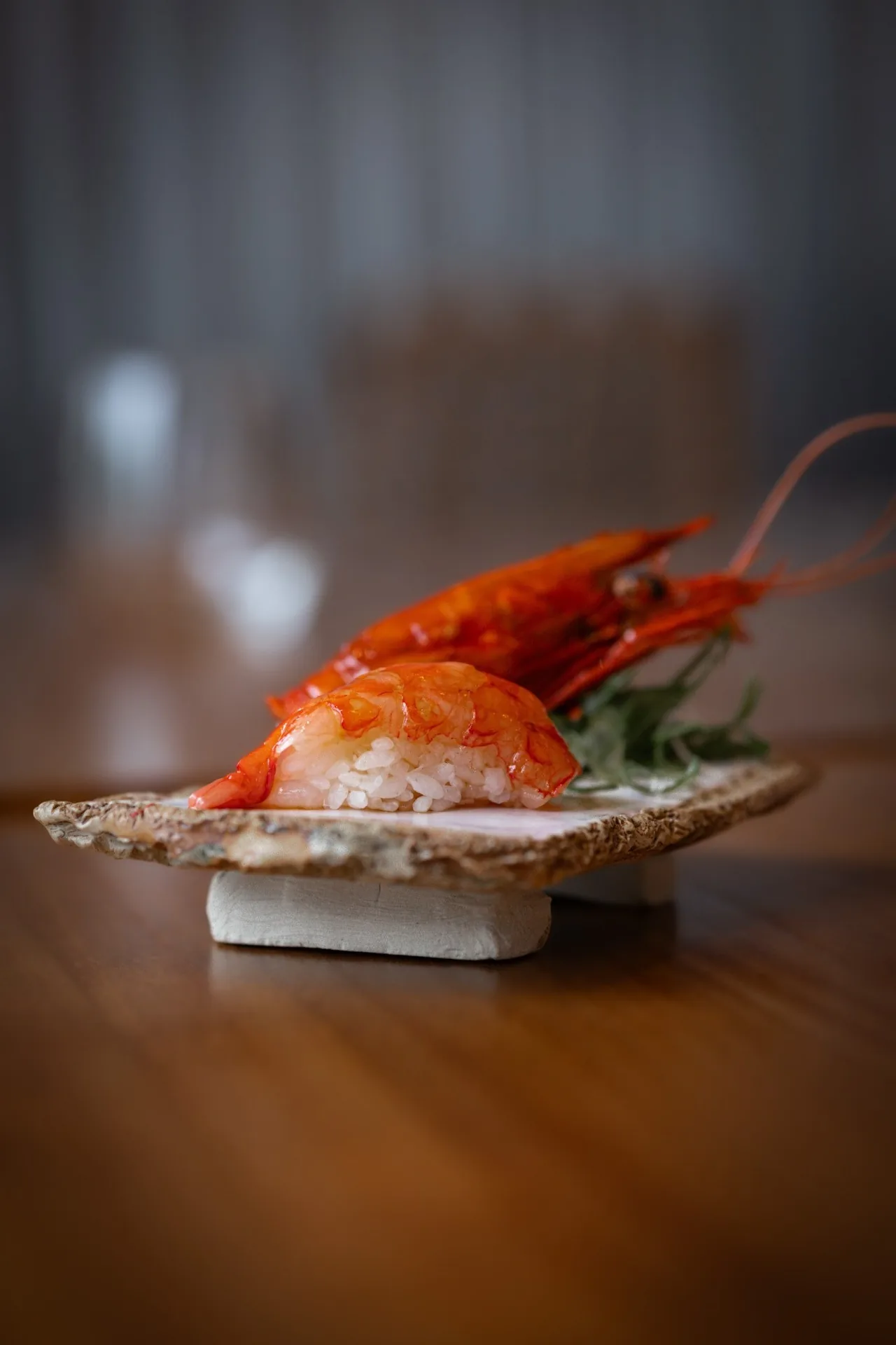 A red prawn nigiri on a small ceramic plate with a whole prawn behind it