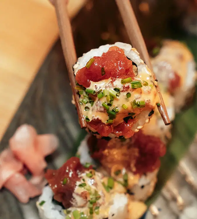 Sushi roll topped with diced tuna, spicy mayo and chives lifted with chopsticks over a plate of more rolls