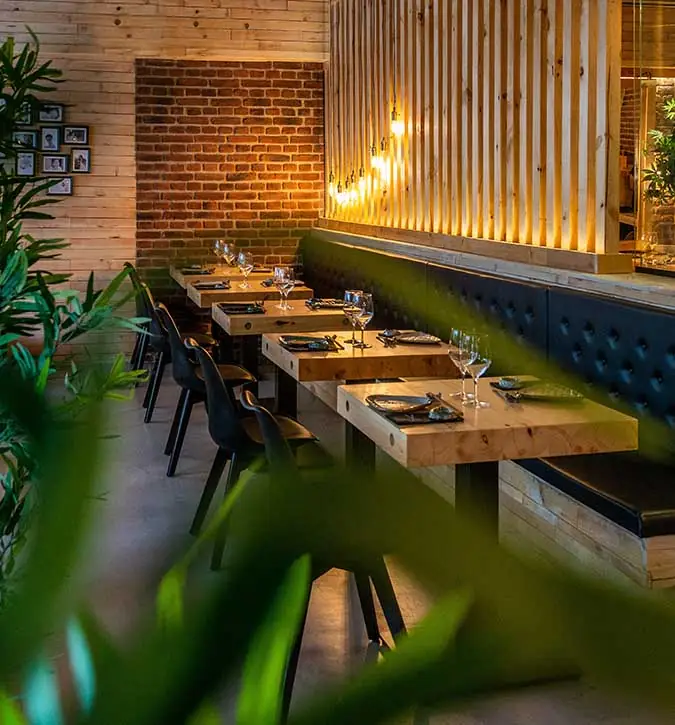 Dining area with wooden tables set with wine glasses along a green tufted banquette, exposed brick and slatted wood wall, plants in foreground