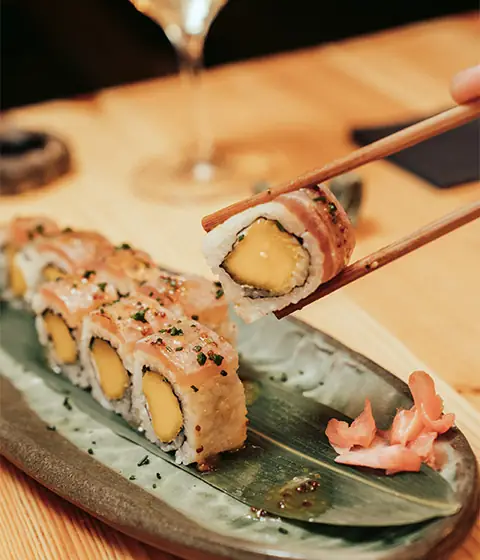 Sushi roll filled with mango and wrapped in seared salmon, a piece lifted with chopsticks, pickled ginger alongside on a green plate