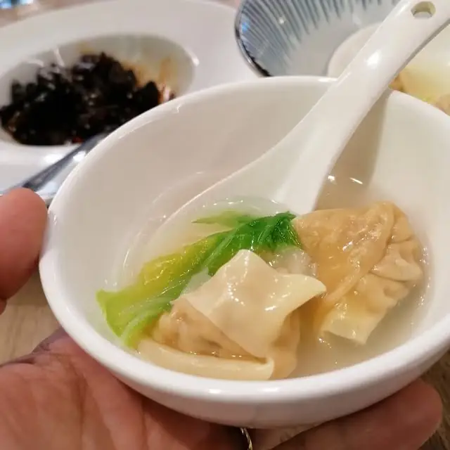 Small white bowl of wonton soup with a dumpling, greens and a soup spoon, held in a hand