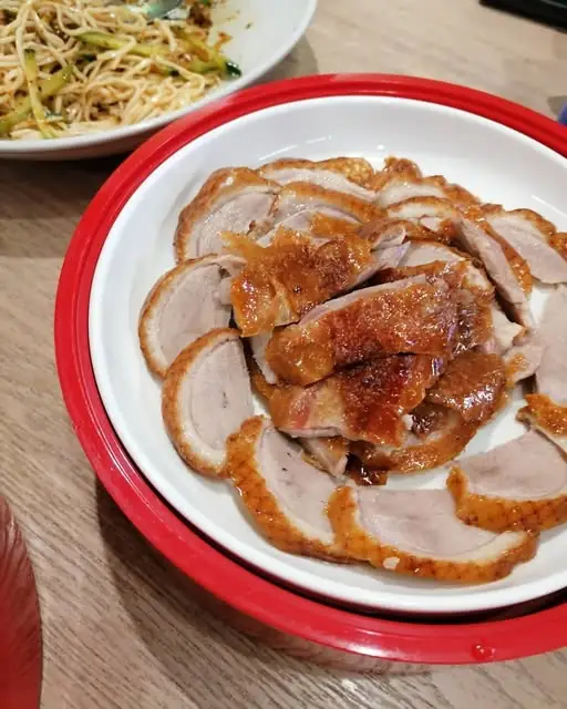 Sliced glazed roast duck in a red-rimmed bowl with a plate of sesame noodles behind on a wooden table