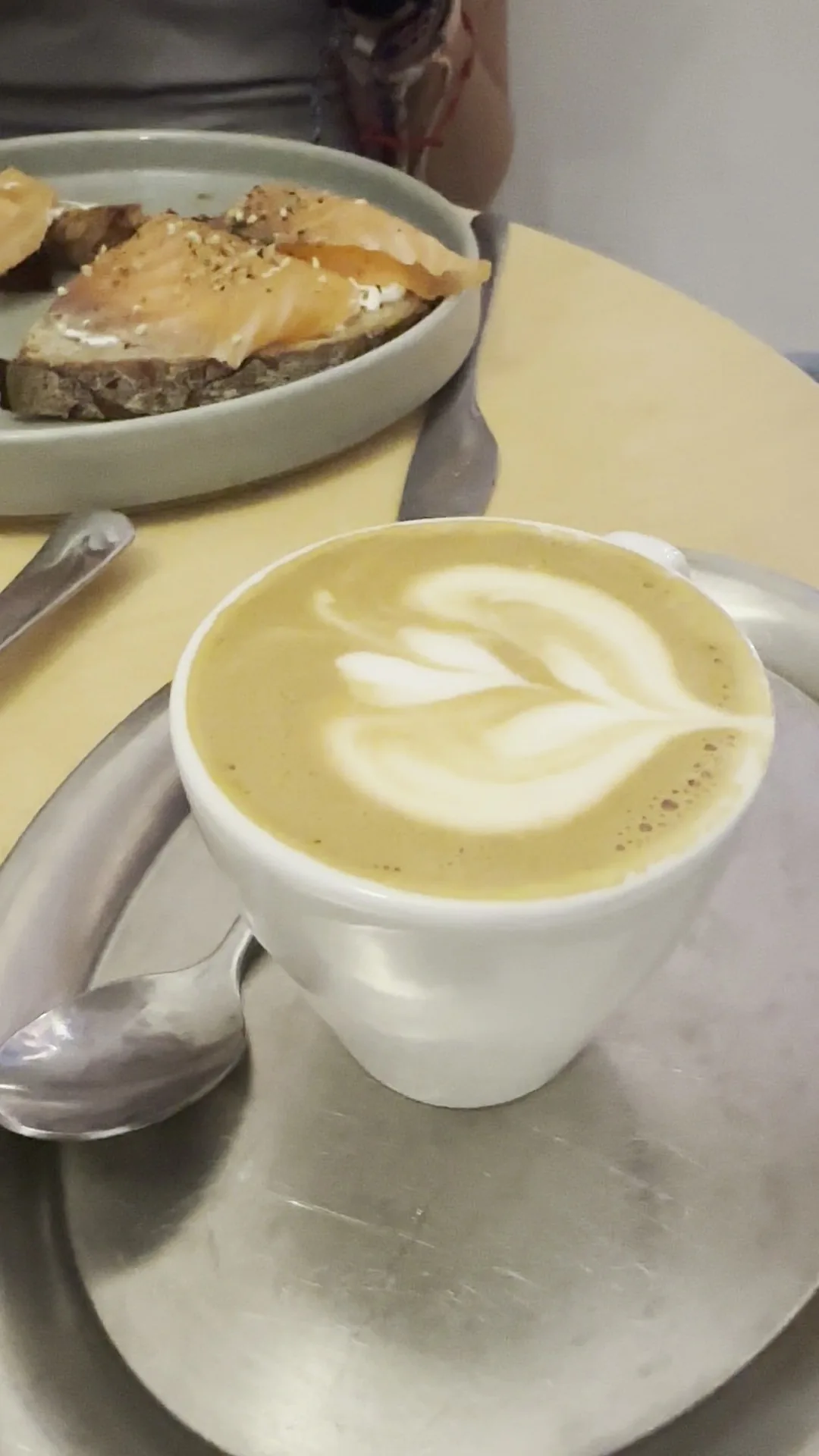 A latte with a leaf pattern in a white cup on a saucer with a spoon