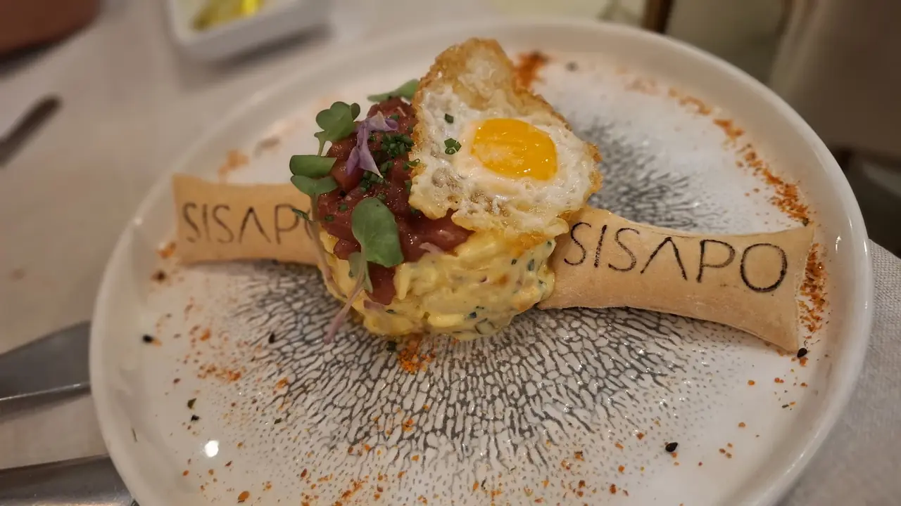 Close-up of a plated dish with fried quail egg, tuna tartare and mashed avocado, flanked by a branded tuile