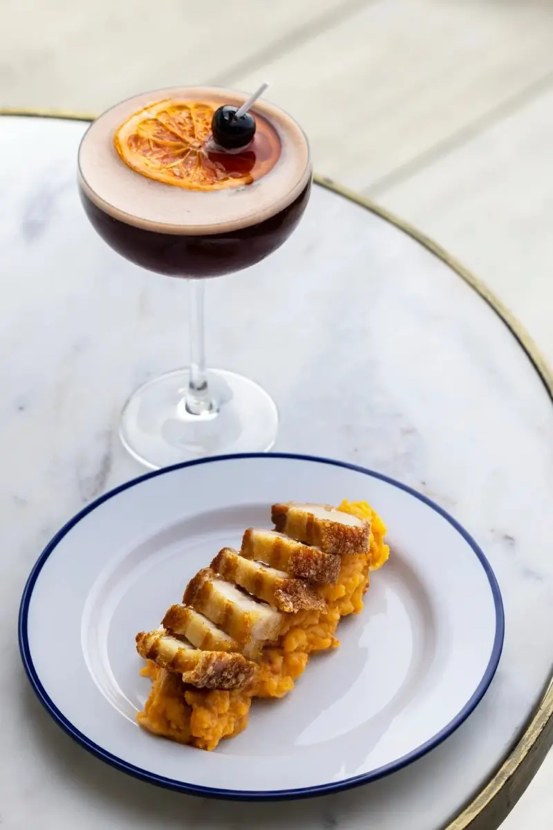 Coupe cocktail with a foamy top garnished with a dried orange wheel and a cherry, beside a white blue-rimmed plate of sliced crispy pork belly over mashed sweet potato on a marble table