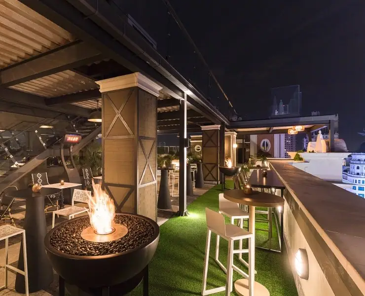 Night rooftop terrace with a lit fire pit, artificial grass, high tables and bar stools, city skyline in the background