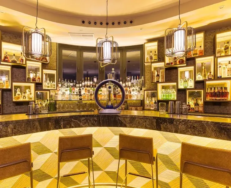 Curved indoor bar with a back wall of illuminated liquor bottles in framed niches, caged pendant lamps and upholstered bar stools