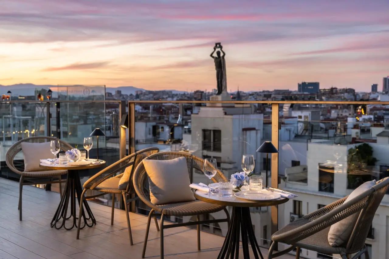 Rooftop terrace set with laid tables and woven chairs overlooking the Madrid skyline at sunset, a rooftop statue and Hotel Emperador glass panel visible