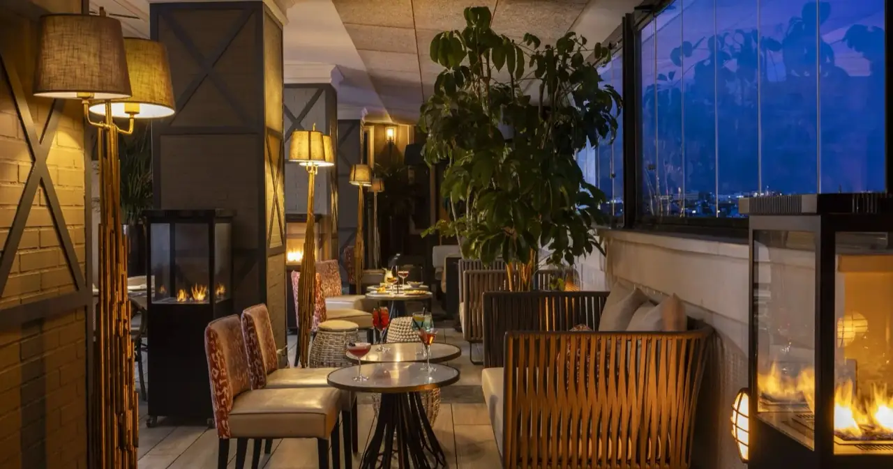 Enclosed terrace lounge at dusk with fireplaces, floor lamps, a large plant and cocktails on marble tables