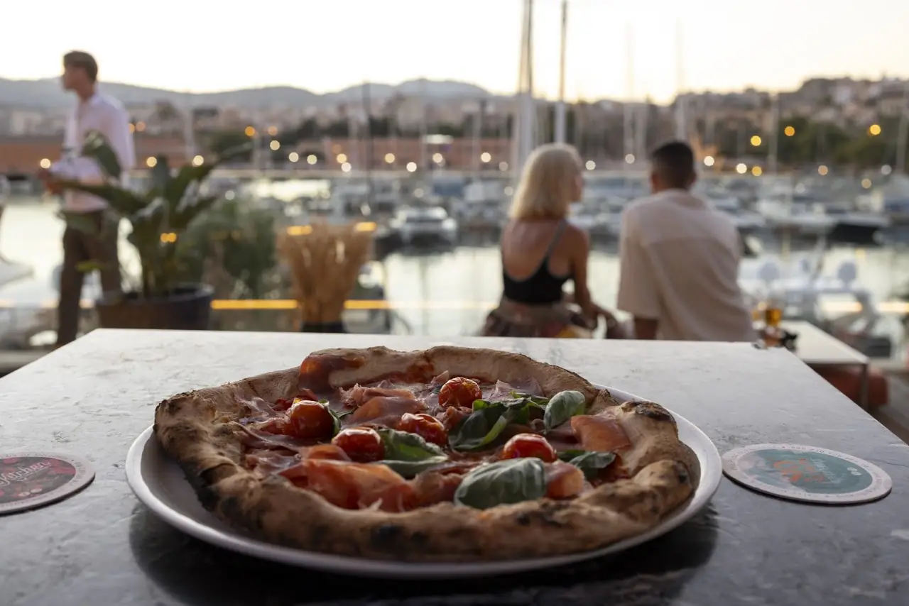 Wood-fired pizza with tomato, mozzarella, and basil on an outdoor table overlooking the marina